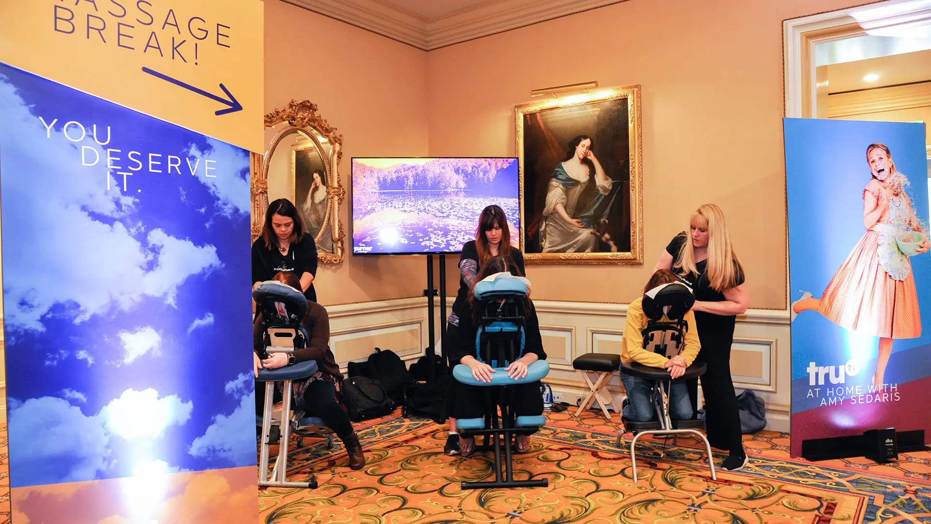 A smiling woman provides a relaxing seated massage to a client using a massage chair. The indoor setting boasts elegant decor and a large mirror on the wall, with a television screen partially visible&mdash;a perfect scene for a rejuvenating Corporate Chair Massage LA experience.