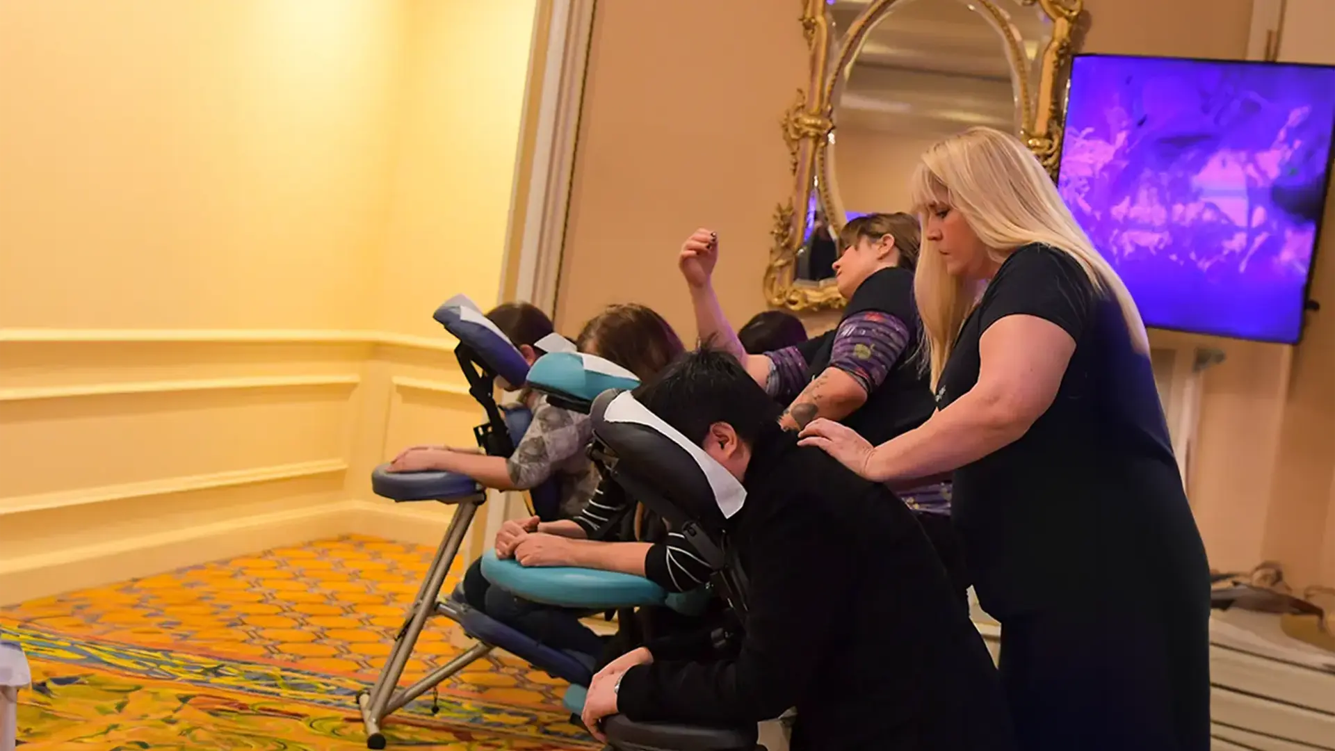 A smiling woman provides a relaxing seated massage to a client using a massage chair. The indoor setting boasts elegant decor and a large mirror on the wall, with a television screen partially visible&mdash;a perfect scene for a rejuvenating Corporate Chair Massage LA experience.