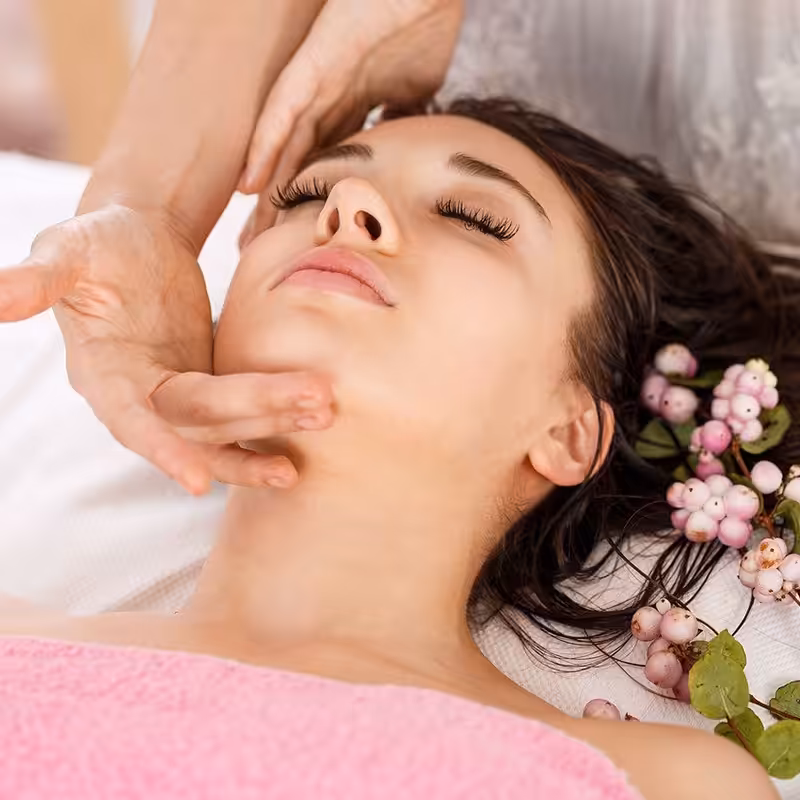 A woman receiving a relaxing facial massage while lying down with eyes closed. Her head is adorned with small pink and white flowers, resting on a white towel. A pink towel covers her shoulders.