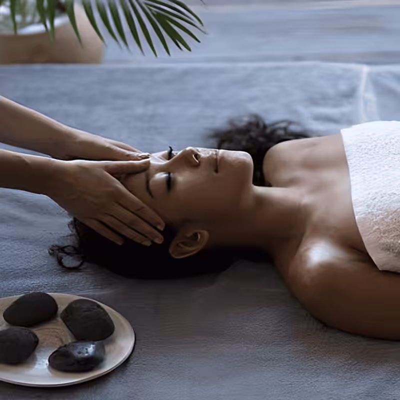 A woman receiving a relaxing facial massage while lying down with eyes closed. Her head is adorned with small pink and white flowers, resting on a white towel. A pink towel covers her shoulders.