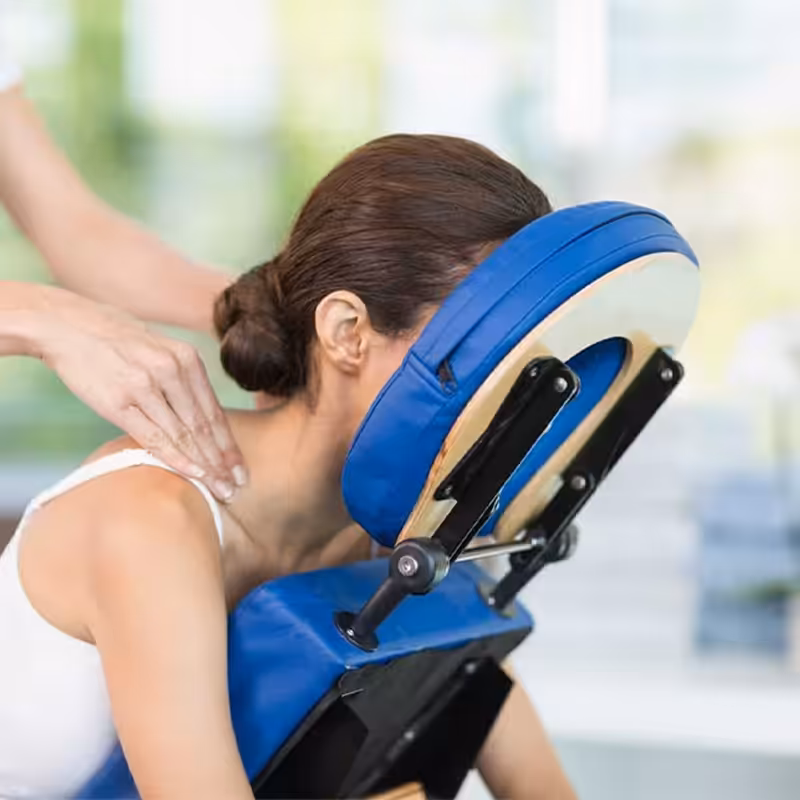A woman receiving a relaxing facial massage while lying down with eyes closed. Her head is adorned with small pink and white flowers, resting on a white towel. A pink towel covers her shoulders.