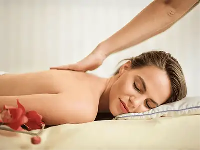 A woman receiving a relaxing facial massage from ACM mobile massage services while lying down with eyes closed. Her head is adorned with small pink and white flowers, resting on a white towel. A pink towel covers her shoulders.